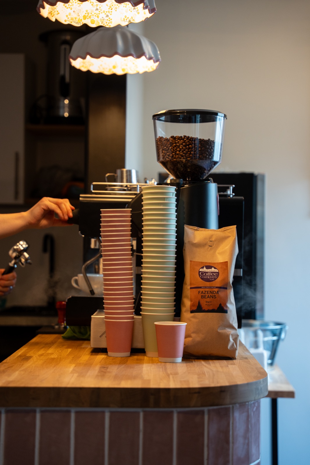 The coffee bar at Blossom cafe in Banbury: espresso machine, grinder full of beans, stacked pink and sage takeaway cups, and a bag of Oxford Coffee Company Fazenda beans on the oak counter